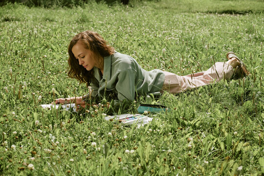 Caucasian young adult woman lying on green grass and drawing in sketchbook during outdoor art master class. Image suitable for promoting creative hobbies and mental wellbeing, wide shot