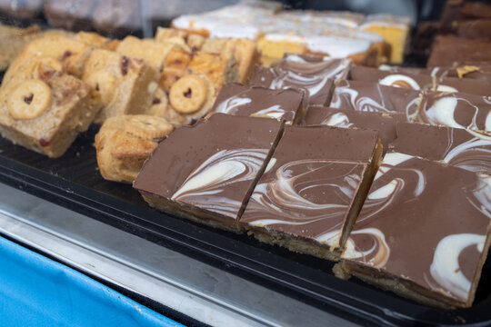 Close up of chocolate topped traybake slices with decorative white chocolate swirls displayed beside biscuit topped cakes and iced sponge squares behind a glass counter at a traditional food stall