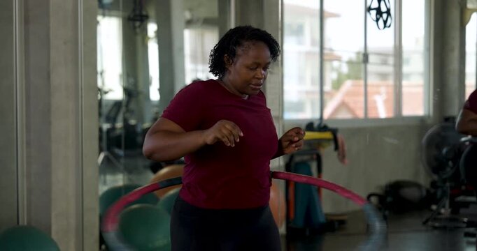African American plus size woman rotates hula hoop around waist in modern gym, moving hips with steady rhythm. Curvy female athlete practices cardio, core balance, confidence, body positivity.