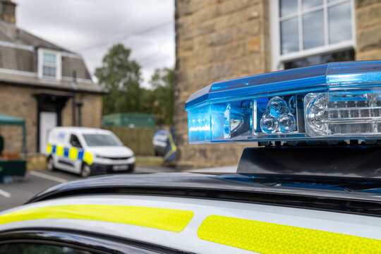 Close up of a blue emergency light bar mounted on the roof of a British police vehicle with high visibility yellow markings and another marked police van softly blurred in the background