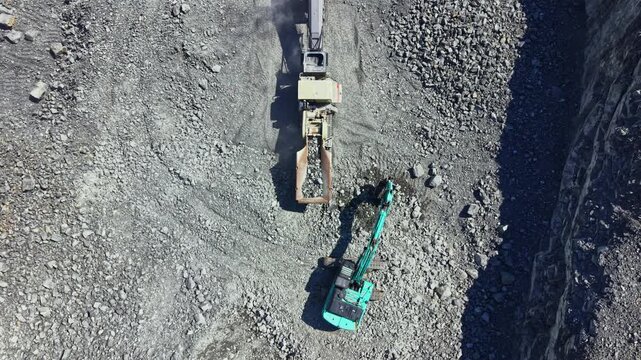 Heavy machinery extracts granite in a quarry. Workers oversee the process as rocks break apart. Dust and gravel cover the ground. This scene captures the activity at the granite site.