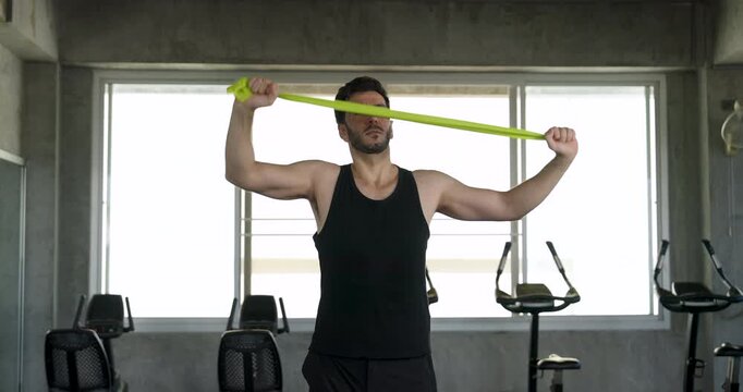 Adult male athlete pulls green resistance band across chest during upper body strength training in modern gym. Shoulder mobility workout builds stability, endurance, flexibility, healthy lifestyle.