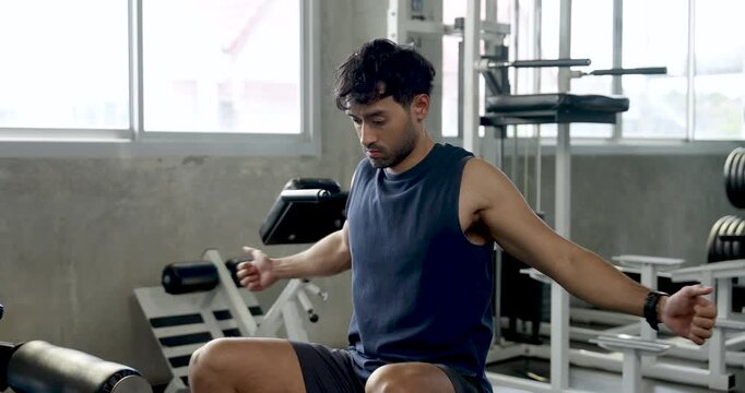 Young Asian man pauses on seated cable row machine in modern gym, raising arms and shaking out tension between sets, solo fitness recovery, arm workout routine, wellness, healthy active lifestyle.