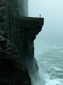 Lone figure stands on precarious cliff edge overlooking turbulent, misty ocean