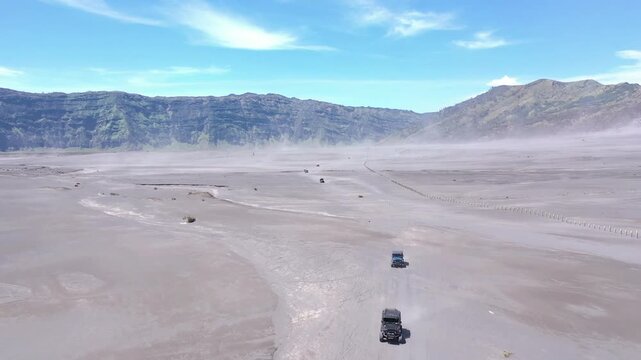 Wide aerial drone view of 4x4 offroad jeeps parked on the black sand sea of Mount Bromo with crater background under blue sky, East Java, Indonesia