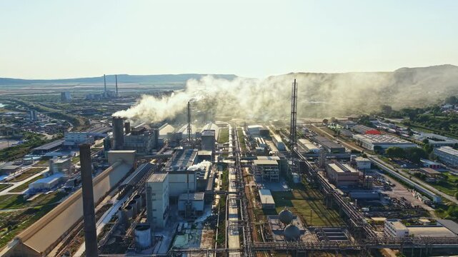 A site shows large structures and machinery in operation under blue skies. Smoke rises from chimneys, and the area is busy. The landscape has many production elements.