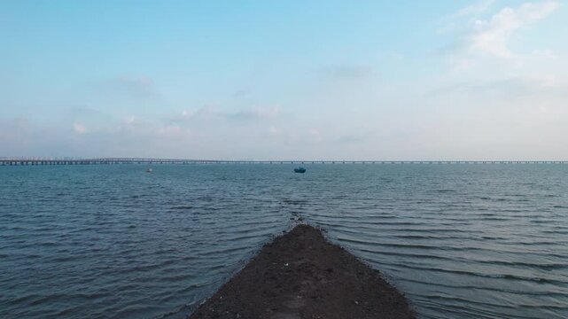 Weathered fishing boats rest on a muddy tidal flat beside calm coastal waters, with a distant bridge under a clear sky.