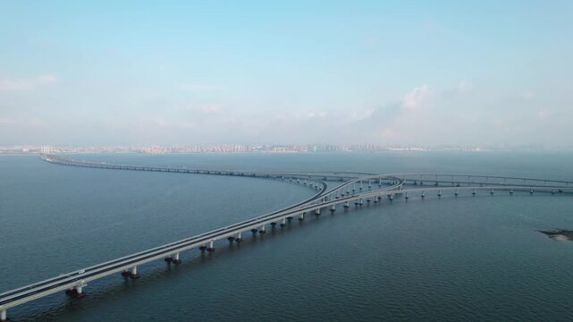 A long curved sea-crossing bridge with multiple road lanes extends over calm blue water towards a distant urban skyline.