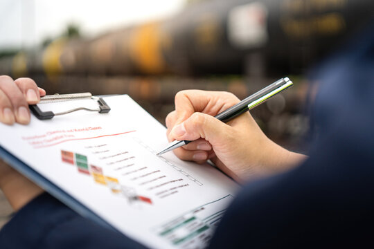 A safety officer is checking on hazardous chemical material checklist for risk assessment before tranportation the dangerous good with train tanker railway background. Industrail working scene.
