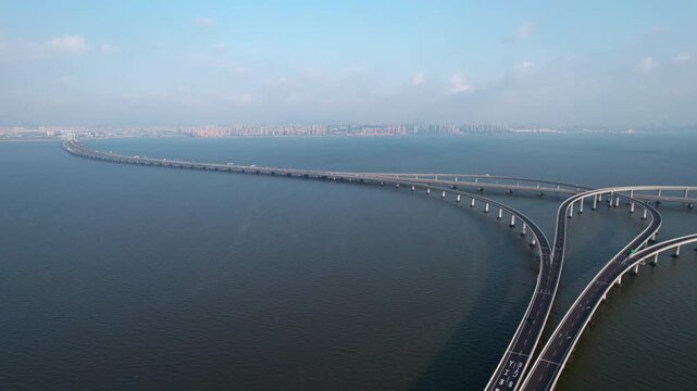 A long concrete coastal bridge spans across a vast blue water body, with road curves and distant city views under a clear sky.