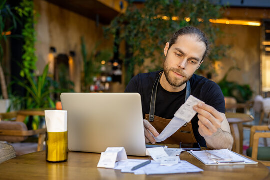 Young barista analyzing receipts near laptop