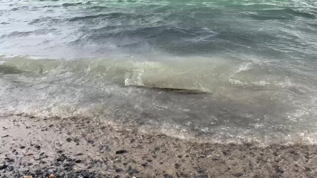 A close-up, ground-level view captures a small, foamy wave actively breaking onto a pebble beach of a lake or ocean under an overcast sky. The image highlights the rough texture of the dark. 