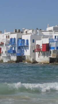 Vertical view of Little Venice in Mykonos Town, featuring waterfront buildings, traditional Cycladic architecture, and the beautiful Aegean Sea coastline.