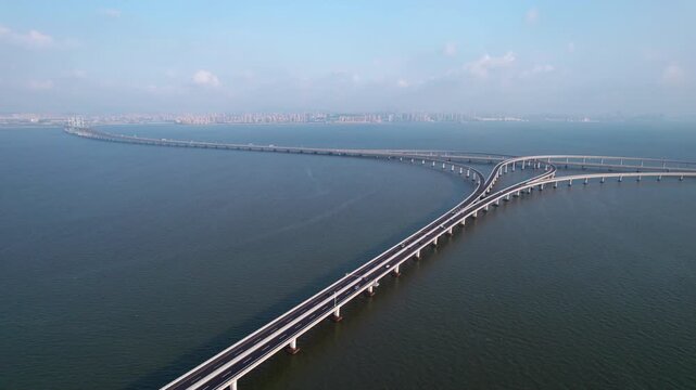 Aerial view of a long sea-crossing bridge with interchange over calm blue water, distant urban skyline on horizon.