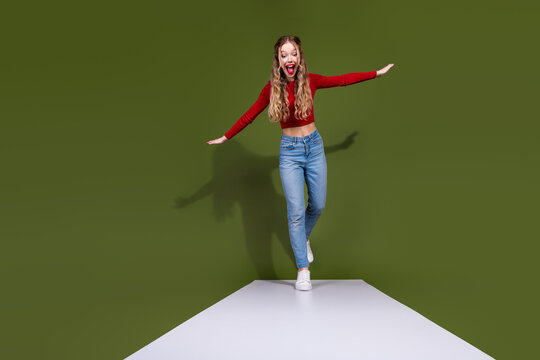 Young woman in red top and jeans walks a white runway against green background with excited smile