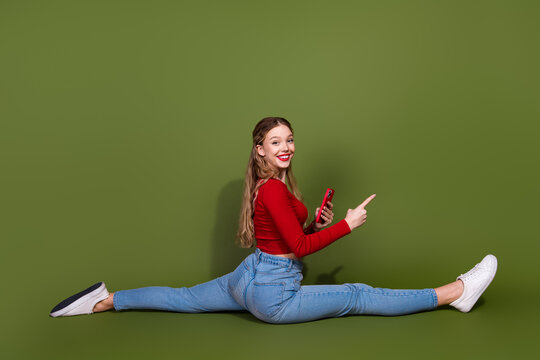 Young woman performing a full split in red top jeans and white sneakers holding a phone smiling against a green background