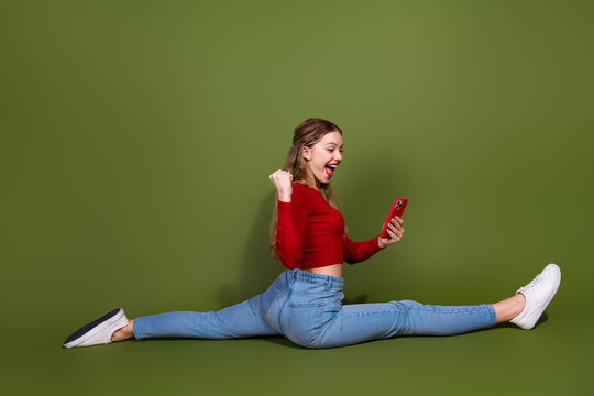 Young woman performs split on green background while using red smartphone joyfully