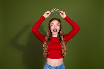 Young woman in red crop top places tiara on head with big smile outdoors on green background © deagreez