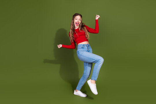 Young woman jumps with joy in red top and jeans against green studio background for lifestyle fashion advertising use