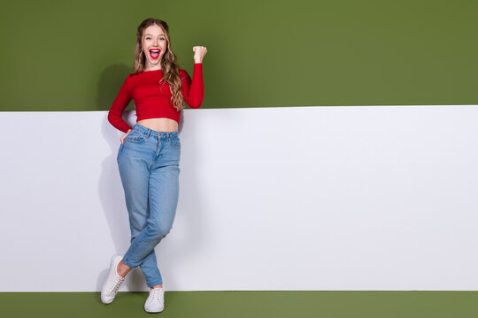Young woman in red crop top and jeans smiles and cheers standing against green white backdrop