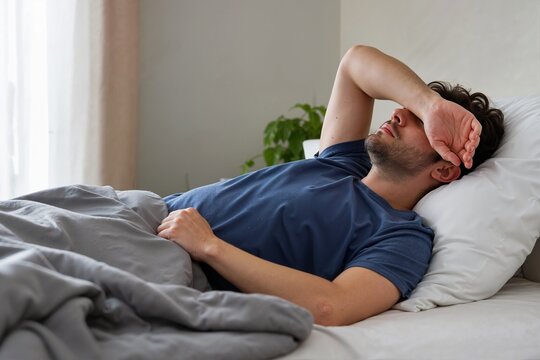 Tired man lying in bed with his arm covering his eyes. Exhausted male patient suffering from a morning headache and hangover. Illness and physical discomfort concept