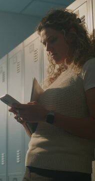 Female Teacher Standing Alone in Empty School Locker Room, Checking Smartphone Notifications, Responding to Messages. Modern Education, Digital Communication, Everyday Teacher Workflow. Vertical Shot.