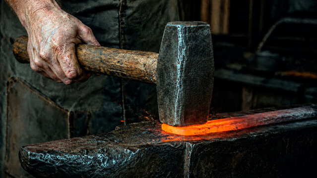 Close-up of a blacksmith striking glowing hot metal with a hammer on an anvil. Traditional metalworking and forging craftsmanship in a dark workshop