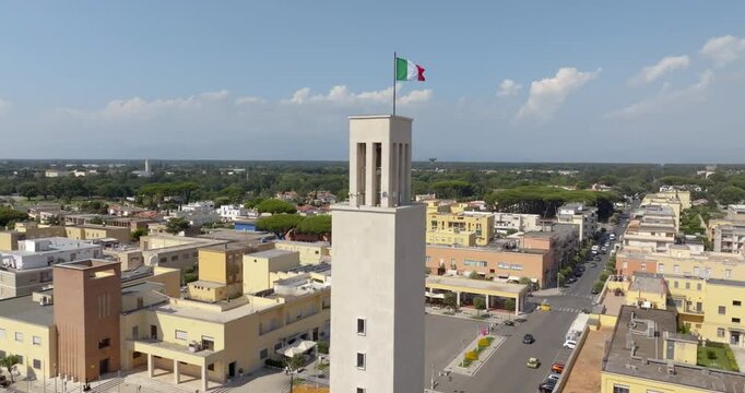 Aerial view of the civic tower of the town hall of Sabaudia, in the province of Latina, in Lazio, Italy, with the Italian flag on the top and the rationalist town centre below, under a blue sky.