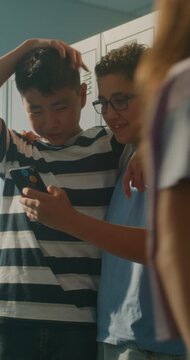 Two Cheerful Teenage Classmates Stand by School Lockers, Sharing Smartphone and Laughing Together Before Class. Authentic Gen Z Lifestyle, Friendship and Positive Social Interaction. Vertical Shot.