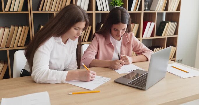 Girls students studying writing homework notes. Two learners work at a desk in a library classroom, taking notes near a laptop while preparing for exams. Concept of teamwork and learning.