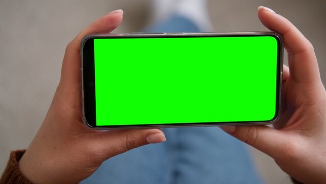 Top view of woman hands holding smartphone horizontally with green screen mockup chroma key blank display. Female user looking at phone template mockup screen for video streaming or mobile gaming