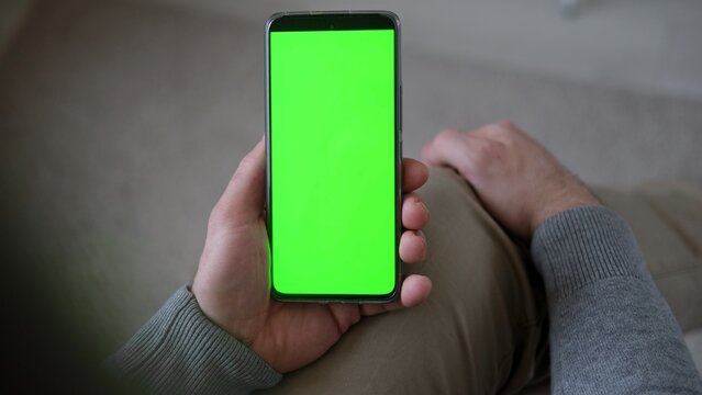 Top view of man holding smartphone vertically with green screen mockup chroma key blank display. Male user sitting and looking at mobile phone template mockup surface inside cozy room at home