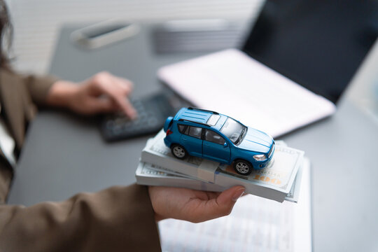 Woman holding car model on stacks of money while using a calculator, representing car loan approval, vehicle financing, credit assessment, purchase payment, budgeting, banking, and financial planning.