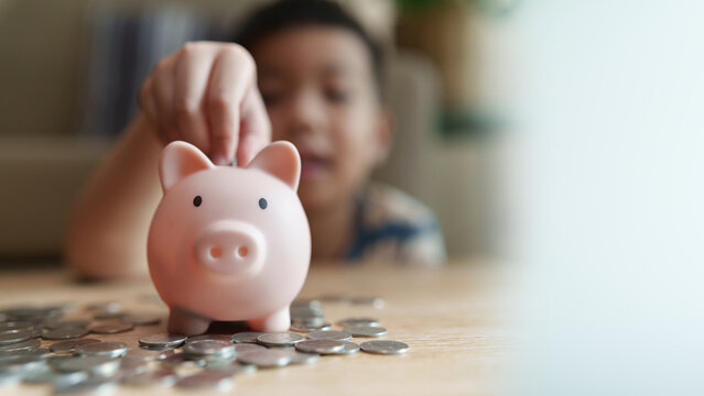 Young Asian boy putting a coin into a piggy bank surrounded by coins, representing money saving, financial education, budgeting, savings goals, future planning with panoramic and copy space.