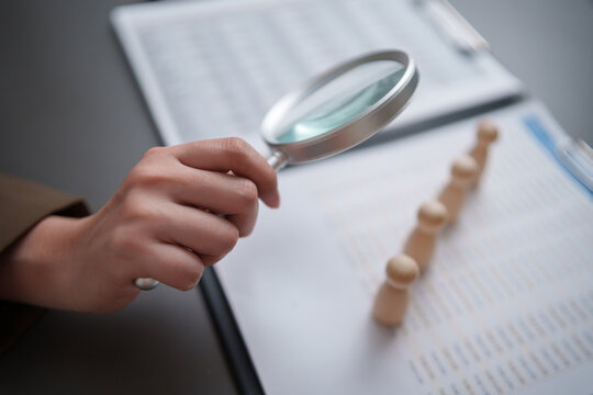 Recruiter using magnifying glass to examine wooden candidate figures and documents, representing recruitment, applicant screening, employee evaluation, talent search, hiring decisions, and HR review.