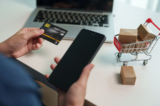 Person holding credit card and smartphone beside shopping cart with parcels and laptop, representing online shopping, e-commerce, digital payment, checkout, product delivery and cashless purchasing.