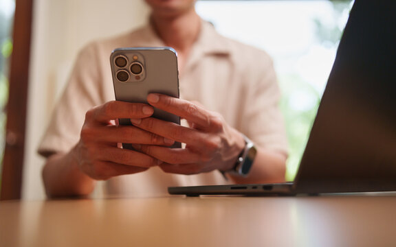 Man using smartphone beside laptop at home, representing mobile browsing, online communication, messaging, digital lifestyle, remote work, internet connectivity, multitasking, and modern technology.
