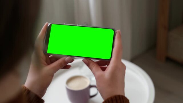 Girl sitting at home and watching movie on mobile phone template mockup with green screen chroma key. Female hands holding smartphone horizontally for digital entertainment leisure in cozy room