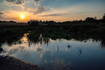 Fototapeta premium Low setting sun glowing through scattered clouds, casting warm golden reflections across a calm wetland pond surrounded by reeds, aquatic grasses, and silhouetted countryside vegetation at dusk.