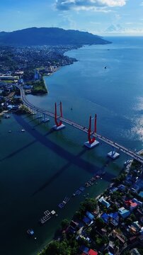 Aerial view of the Merah Putih Bridge spanning the bay with its distinctive red towers, surrounded by coastal urban areas and lush green hills in Kota Ambon, Maluku, Indonesia.