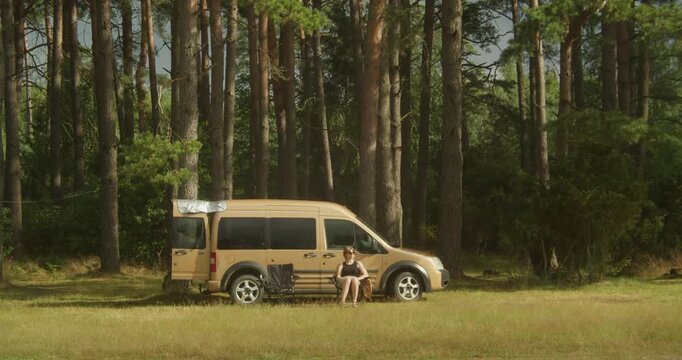 An authentic scene of a digital nomad sitting in a chair outside her camper van in a sunlit pine forest. Perfect for themes of van life, sustainability, and remote work.