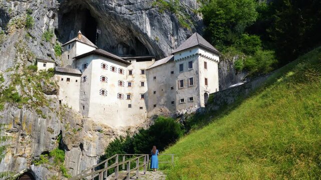 Woman In Blue Dress Stands At Viewpoint Overlooking Predjama Castle In Slovenia. Aerial Rotate Shot