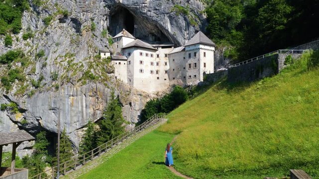 Woman Walks Along Trail In Front Of The Historic Predjama Castle In Slovenia. Aerial Drone Shot
