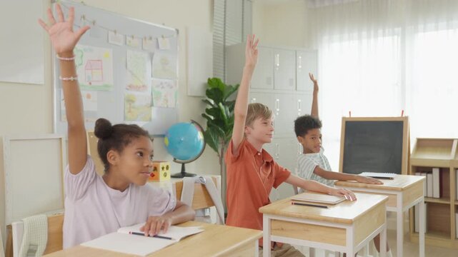 Elementary classroom diverse children learning at wooden desk, writing in notebook, raised hand near globe, chalkboard, window light, colorful posters, engaged school lesson
