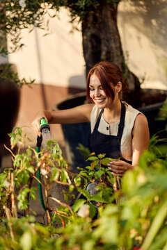 Smiling Woman Watering Plants in Backyard Garden Wearing Apron Enjoying Urban Gardening and Plant Care