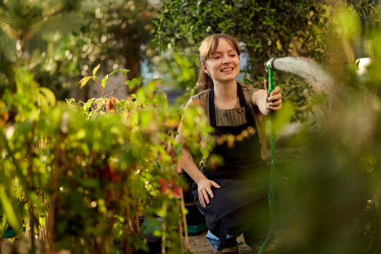 Young Woman Watering Plants in Greenhouse Garden Wearing Apron and Smiling While Gardening