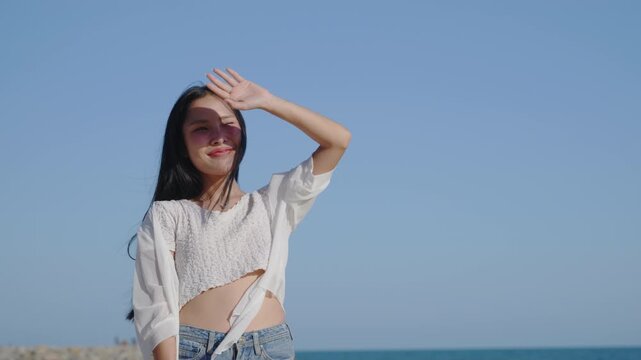 Happy woman on summer vacation beach, shielding eyes from sun while smiling happily under clear blue sky, wearing white casual clothes with long dark hair blowing in gentle sea breeze.