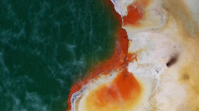 Top down aerial close up of colorful geothermal hot spring with emerald water bordered by vivid orange mineral deposits in Rotorua, Bay of Plenty, North Island, New Zealand, abstract natural patterns