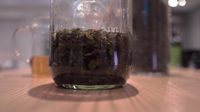 Close-up shot of a large glass jar filled with steeped tea leaves being set down on a wooden table next to a glass beaker and dried tea container.