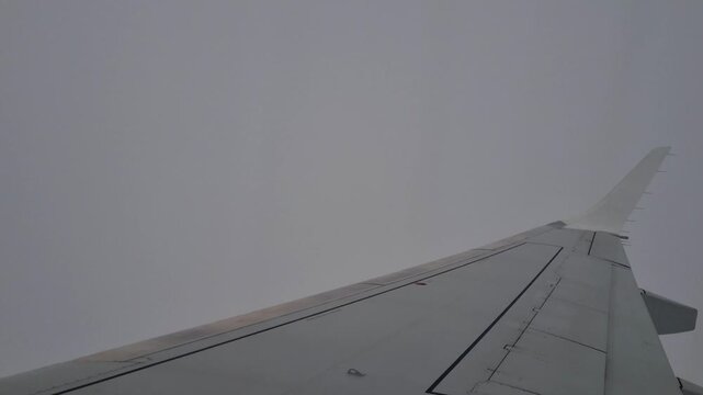 Airplane window view of the wing cutting through thick grey clouds during turbulent flight, capturing a dramatic travel experience and commercial aviation journey above the clouds